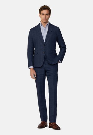 Marineblauw pak van textuurstof, met een enkelvoudige blazer met geknopt revers en slim-fit broek. Bruine leren schoenen.