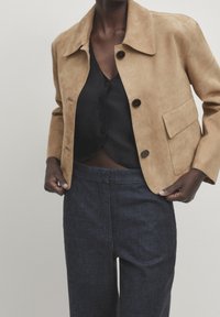 Veste en daim beige avec col, fermetures à boutons et poches latérales, portée sur un haut noir boutonné et un pantalon en jean foncé.