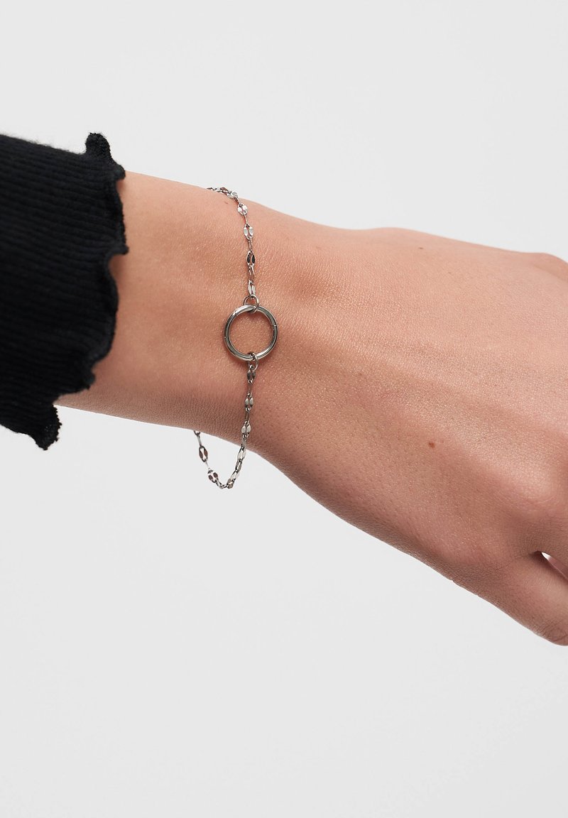 Silver metal bracelet featuring a circular charm, elongated link chain, and a textured surface, worn on a wrist against a neutral background.
