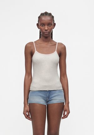 Žena s vrkočmi, ktorá má na sebe svetlobéžové tielko s tenkými ramienkami a modré denimové šortky, stojaca pred jednoduchým bielym pozadím.