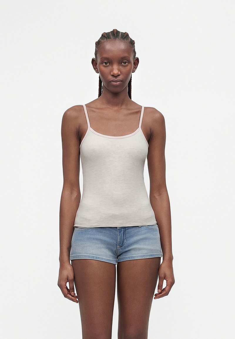 Γυναίκα με πλεξούδες, που φοράει ανοιχτό μπεζ τοπ με λεπτές τιράντες και μπλε τζιν σορτς, στέκεται μπροστά σε ένα απλό λευκό φόντο.