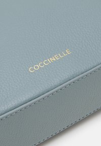 Jasnoniebieska skórzana kopertówka o fakturze, z złotym logo "Coccinelle", ozdobiona przeszyciem na krawędziach i gładkim wykończeniem.