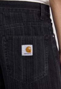 Zwarte denim jeans met een achterzak met een wit label waarop een geel logo en de tekst "carhartt" staat. Verticale streeptextuur zichtbaar.