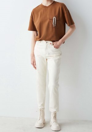 Person trägt ein braunes Kurzarmoberteil mit dekorativer Brosche an der Brusttasche, weiße High-Waist-Jeans und cremefarbene Schnürstiefel.