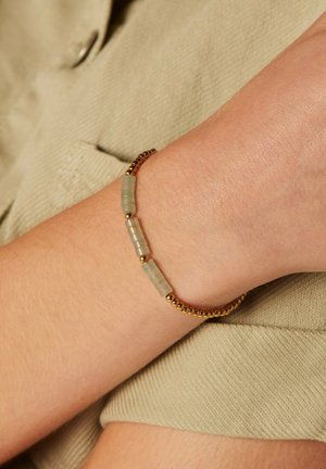 Goudkleurige armband met groene rechthoekige kralen, een gladde textuur en kleine bolvormige accenten. Draagt op een pols tegen een stoffen achtergrond.
