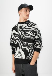Jonge man met een zwarte pet, een zwart-wit abstract patroon trui en donkere jeans, staand met één hand in zijn zak.