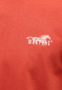 Graphisme d’un cheval mustang blanc et texte « MUSTANG » imprimés sur le haut de la poitrine d’un vêtement en tissu rouge-orange.