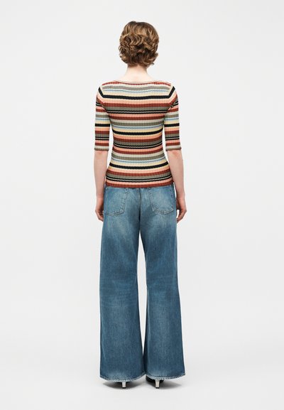 Γυναίκα με κοντά σγουρά μαλλιά, ντυμένη με εφαρμοστό, ριγέ πουκάμισο και φαρδιά μπλε τζιν παντελόνια, που στέκεται μπροστά σε ανοιχτόχρωμο απλό φόντο.