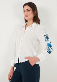 Chemise en lin blanche avec un devant à boutons et des manches longues ; présente des broderies florales bleues sur la manche gauche. Portée avec un pantalon marine.