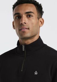 Schwarzer Pullover mit Reißverschluss aus weichem Material. Er verfügt über einen Stehkragen und ein weißes Logo eines Pinguins auf der linken Brustseite.