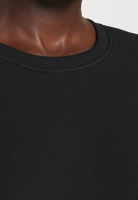 Schwarzer Rundhalsausschnitt-Pullover mit geribbeltem Kragen und strukturiertem Stoff, die Nahaufnahme des Ausschnitts und des Schulterbereichs.