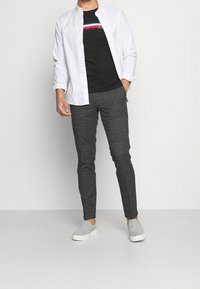 Weißes Hemd über einem schwarzen T-Shirt mit bunten Streifen, kombiniert mit grauen Karo-Hosen und grauen Slippern.