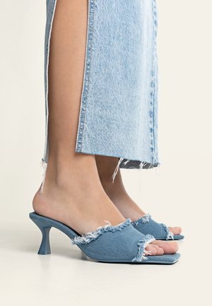 Füße in blauen Denim Open-Toe Mule-Heels mit ausgefransten Kanten, kombiniert mit hellblauen Denim-Cropped-Jeans mit Seitenschlitzen.