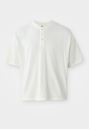 Camisa blanca de manga corta con escote tipo henley, que cuenta con tres botones y un pequeño logo en el lado izquierdo del pecho. Fabricada en tela de algodón.