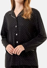 Camisa de pijama negra con bolitas blancas, de mangas largas y con cierre de botones en la parte delantera. Tiene un cuello con muesca y dos bolsillos laterales.