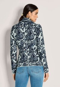 Eine Frau mit dunklem Haar im Pferdeschwanz trägt einen blau gemusterten Pullover im Paisleydesign und hellblaue Jeans, seitlich vor einem schlichten Hintergrund stehend.