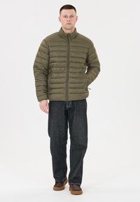 Olivgrön pufferjacka med horisontell sömnad, dragkedjestängning och ståkrage. Bärs med mörka denimjeans och bruna skor.