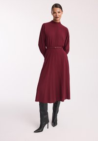 Bordowa sukienka z długim rękawem, wysokim kołnierzem i paskiem z detalami guzików, zestawiona z czarnymi wysokimi botkami na szpilce.