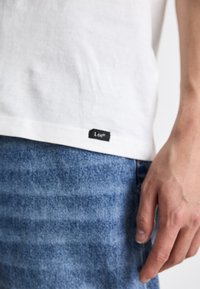 Hvit bomulls t-skjorte med svart "Lee" merkelapp i sømmen, paret med blå denimbukser med en falmet tekstur.