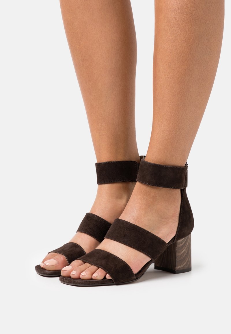 Jambes portant des sandales à talon bloc en daim brun foncé à bout ouvert avec des brides à la cheville et au pied sur fond blanc.