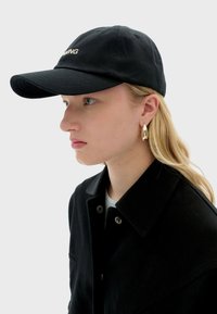Casquette de baseball noire en coton avec une visière incurvée et broderie "RUNNING" en blanc. La personne qui la porte a les cheveux blonds et est vêtue d'une veste noire.