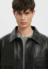 Must nahast jakk klassikalise kraega, nööpidega kinnitusega ja esitaskuga. Kandes halli kampsuni üle, on sellel sile tekstuur.