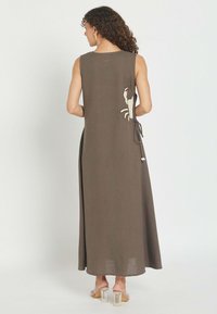 Mujer con cabello rizado que lleva un vestido maxi sin mangas de color verde oliva con un diseño lateral de cangrejo y sandalias transparentes de tacón ancho.