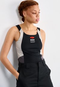 Fekete ujjatlan Nike póló szürke akcentusokkal és vörös swoosh logóval, magas derekú fekete nadrággal párosítva. Simább, sportos szabás.