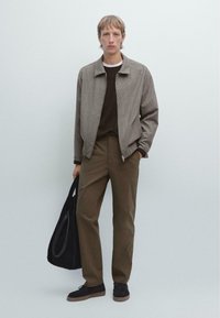 Grijze gestructureerde ritsjack over een bruine trui, olijfgroene broek en zwarte schoenen. Draagt een zwarte tas en staat tegen een lichte achtergrond.