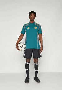 Turkusowy stroj sportowy z żółtymi akcentami, czarne spodenki i czarne skarpety z białymi paskami; zawodnik trzymający piłkę nożną.