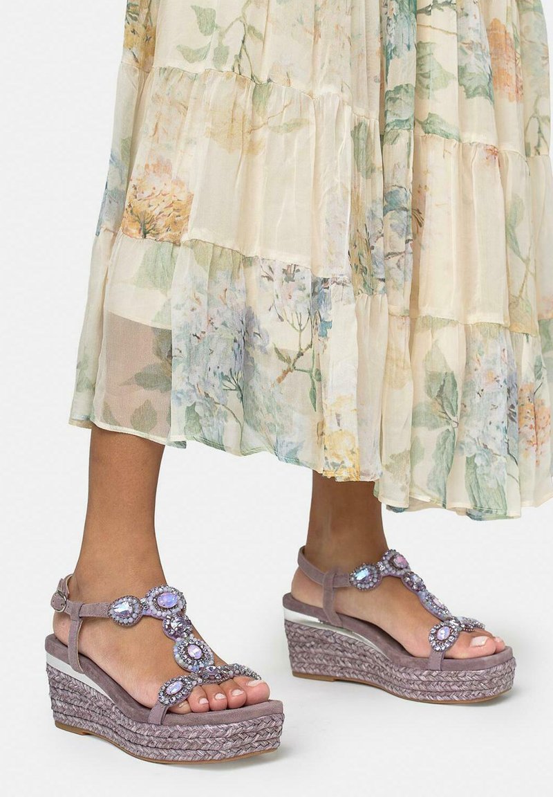 Pieds chaussés de sandales compensées violettes claires ornées de grandes pierres iridescentes, associés à une jupe midi transparente à fleurs aux couleurs pastel douces.