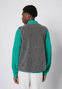 Graue Fleece-Weste mit einer strukturierten Oberfläche, mit einem abgerundeten Halsausschnitt und Armausschnitten, getragen über einem grünen Pullover. Die Weste hat eine lässige Passform.