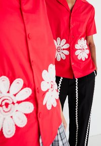 Červená košeľa s krátkymi rukávmi a bielou vyšívanou kvetinovou aplikáciou na prednej strane, s voľným strihom a zapínaním na gombíky.