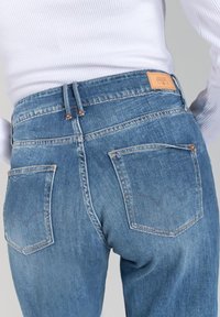 Jean en denim bleu à taille haute, avec deux poches arrière à couture incurvée et une étiquette de marque en cuir sur la ceinture.