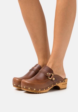 Brown leather clogs with a wooden sole, featuring a strap with a gold buckle and brass studs along the base, open back design.