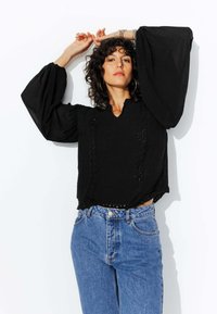 Haut en tricot noir avec des manches larges et bouffantes, présentant une finition texturée et de petits détails en découpes, associé à un jean bleu taille haute.