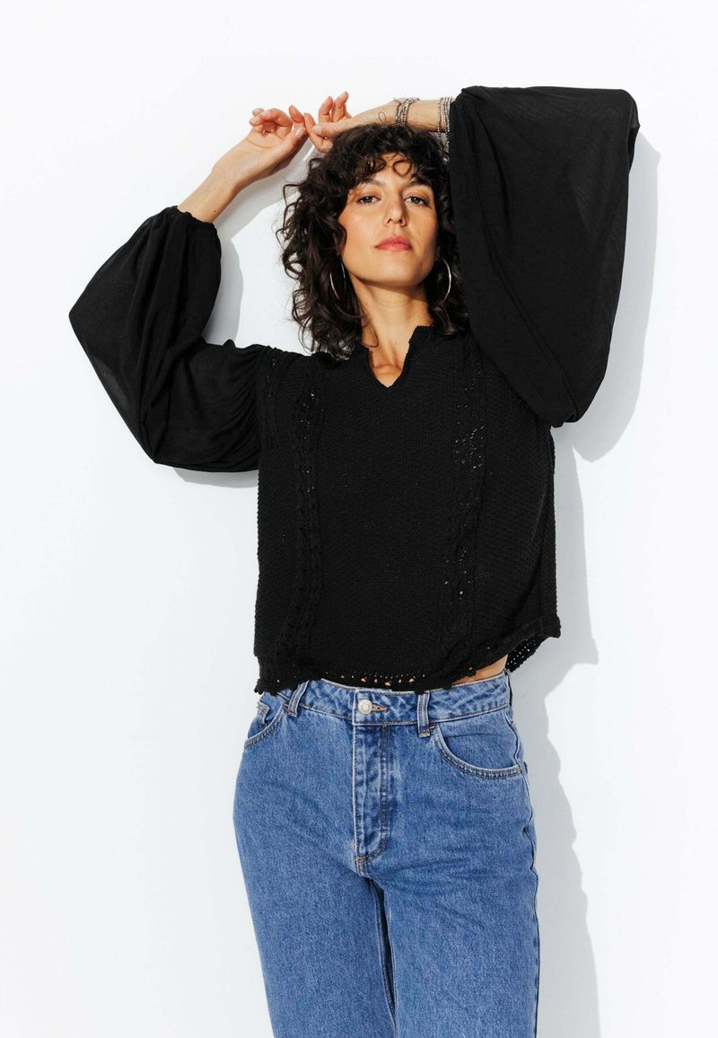 Haut en tricot noir avec des manches larges et bouffantes, présentant une finition texturée et de petits détails en découpes, associé à un jean bleu taille haute.