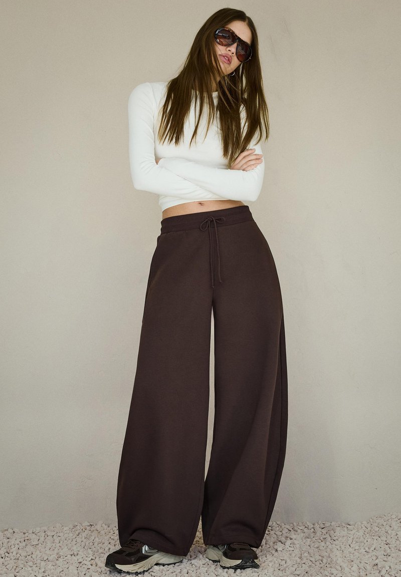 Hnedé široké nohavice so sťahovacím pásom, v kombinácii s priliehavým bielym dlhým rukávom a čiernymi teniskami. Modelka má rovné hnedé vlasy.