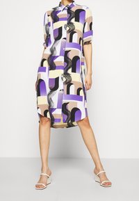Femme portant une robe chemise mi-longue avec des motifs géométriques abstraits en violet, beige, noir et crème, ainsi que des sandales à talons blancs.