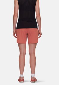Krótkie spodenki sportowe w kolorze koralowym, lekkiego materiału, gładkiej tekstury. Noszone z dopasowanym czarnym topem na ramiączkach i butami do biegania z wzorzystą podeszwą.