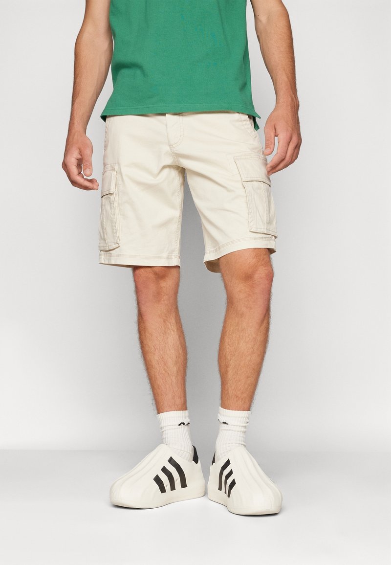 Homme portant un short cargo beige, des chaussettes blanches, des chaussures blanches slip-on avec des rayures noires et une chemise verte sur un fond uni.