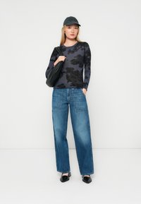 Top ajustado de manga larga en gris oscuro con patrones de nubes negras, combinado con jeans de pierna ancha en azul, tacones cuadrados negros y una gorra oscura.