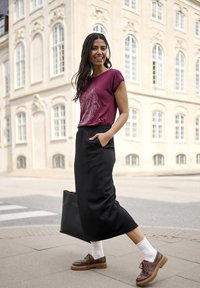 Chemise courte à manches en bordeaux avec un imprimé décoratif, associée à une jupe noire longueur cheville. Chaussures marron à semelles épaisses et chaussettes blanches. Sac fourre-tout noir.