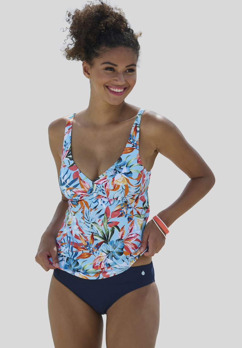 Junge Frau lächelt, trägt einen geblümten Tankini-Oberteil mit blauen, orangefarbenen und grünen Blättern und einfarbige dunkelblaue Bikinihosen.