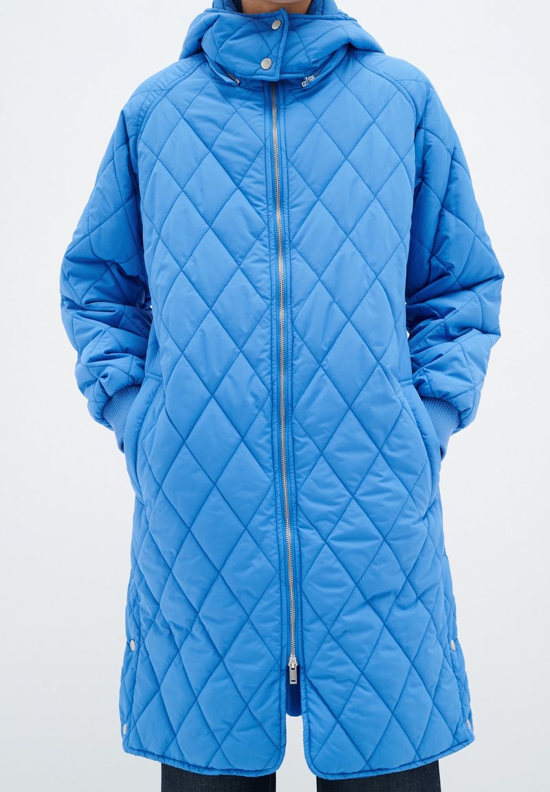 Chaqueta acolchada azul con cremallera, capucha y bolsillos laterales. La tela tiene una textura suave con un patrón de diamantes y puños acanalados.