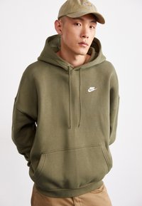 Olīvzaļš kapucīna džemperis ar kangauru kabatu un auklu kapuci. Uz kreisā krūts ir balts Nike logo. Mīksts audums ar izturīgu tekstūru.