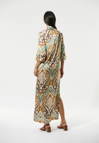 Robe longue à motifs présentant un mélange de teintes dorées, de teal et de marron, avec des manches larges et une fente sur le côté. Tissu lisse et coupe décontractée.