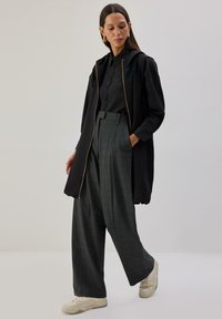 Črna jakna z zadrgo in kapuco, oblečena preko črne srajce s gumbi ter sivih širokih hlač s vzorcem. Kremaste superge z reliefnim podplatom.