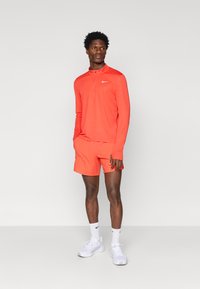 Nike athletic wear featuring a half-zip long-sleeve top and matching shorts in bright orange, with textured fabric and white logo accents.