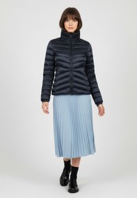 Chaqueta de plumas azul marino con cuello alto y costuras en forma de chevron, combinada con una falda plisada azul claro y zapatos negros brillantes.